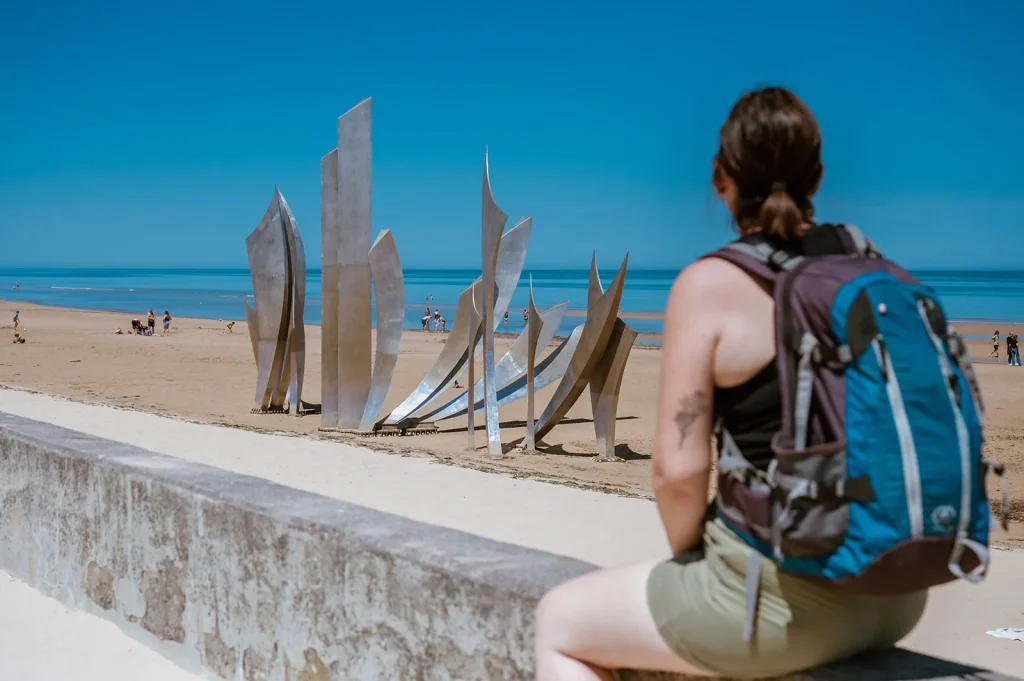 Randonneuse avec un sac à dos regardant la statue Les Braves sur la plage de Saint-Laurent-sur-Mer