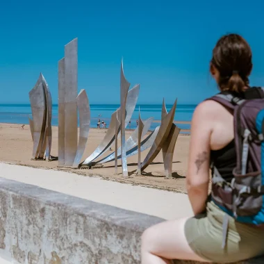 Randonneuse avec un sac à dos regardant la statue Les Braves sur la plage de Saint-Laurent-sur-Mer